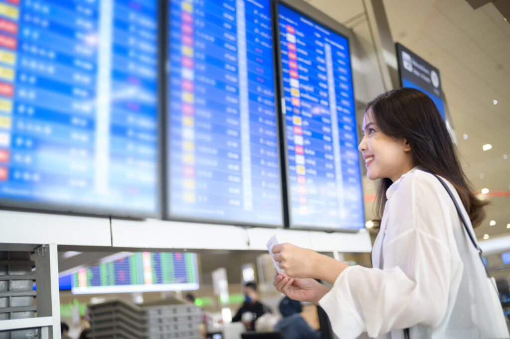 Woman in white shirt smiles while checking large flight-information screens at an airport.